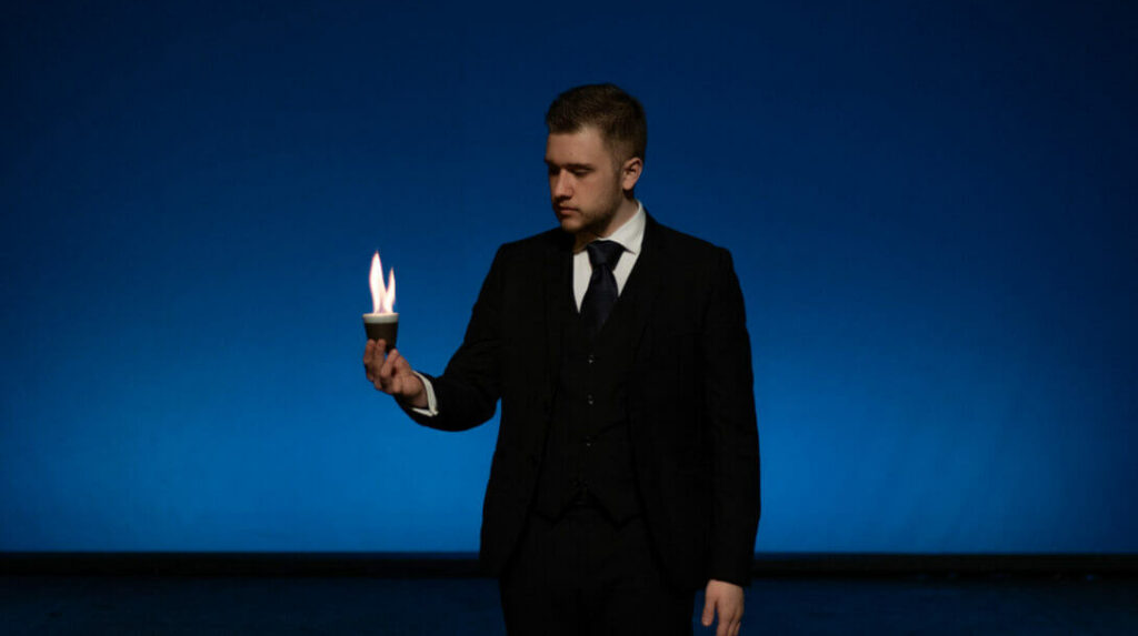 Friedemann steht vor einem tiefblauen Hintergrund, in einem dunklen Anzug mit Krawatte. Er hält eine kleine Espresso-Tasse in seiner rechten Hand, aus der Flammen aufsteigen. Sein Gesichtsausdruck ist ernst und konzentriert, während das blaue Licht den Kontrast zur leuchtenden Flamme verstärkt.